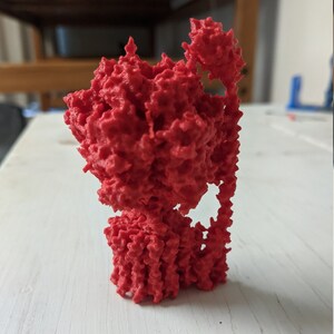 May include: A 3D-printed, bright red, abstract sculpture with a complex, textured surface. The form is irregular, with a dense, porous structure and a vertical orientation. The sculpture appears to be made of a solid material.