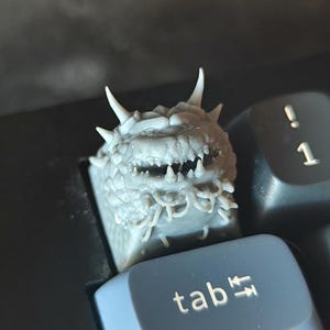 May include: A detailed, gray dragon-themed keycap, featuring sharp teeth, horns, and intricate textures. The keycap is designed to replace a standard keyboard key, shown here on the 'tab' key. The design is a unique accessory for computer enthusiasts.