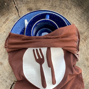 May include: A set of blue speckled camping dishes, including plates, a cup, and a knife, are nestled inside a brown cloth bag. The bag features a white circle with a fork and knife graphic, suggesting a picnic or outdoor dining theme.