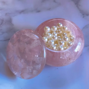 Puede incluir: Una caja de baratijas ovalada rosa con tapa a juego. La caja está llena de perlas blancas. La caja y la tapa tienen una apariencia translúcida, revelando la textura del material.