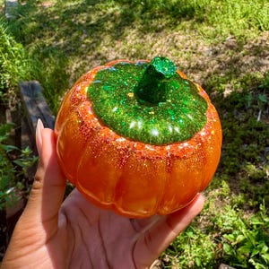 May include: A decorative pumpkin-shaped container with a glittery orange body and a green glitter top. The container is held in a hand against a background of green grass and sunlight.