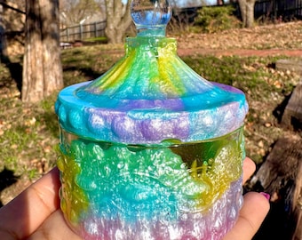 May include: A colorful glass jar with a domed lid. The jar has a tie-dye pattern in shades of blue, green, yellow, and purple. The lid is clear glass with a decorative knob.