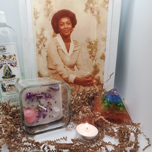 May include: A square glass candle holder with a pink and purple candle inside. The candle has glitter and crystals embedded in it. There is a small, lit tea light candle in front of the holder. A rainbow colored crystal pyramid is to the right of the candle holder. The items are on a brown paper surface.