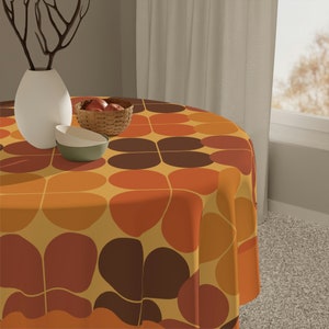 Puede incluir: Un mantel redondo con un patrón retro de círculos superpuestos de color marrón, naranja y amarillo. El mantel está puesto sobre una mesa con un jarrón blanco y un cuenco de fruta.