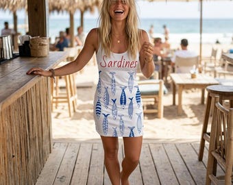 May include: White sleeveless dress with blue fish patterns. The word "Sardines" is written in red across the chest. The dress is knee-length and is worn by a person standing on a wooden deck near a beach.