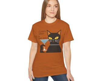 May include: An orange t-shirt featuring a black cat illustration holding a martini glass. The cat has yellow eyes and a white collar. Text on the shirt reads "Classy, Sassy & Bad Assy."