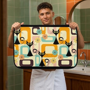 May include: Rectangular bath mat with a retro design featuring black cat silhouettes, geometric shapes, and starbursts in orange, teal, and cream colors. The mat has a black border and is held up by a person.