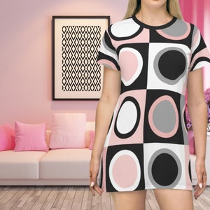 May include: A short-sleeved dress with a geometric pattern of pink, white, gray, and black circles and squares. The dress is worn by a person in a pink room with a white couch and a framed black and white diamond pattern.