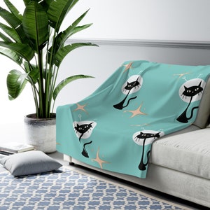 May include: A teal blue throw blanket with a repeating pattern of black cats with antennas and gold stars. The blanket is draped over a white couch.