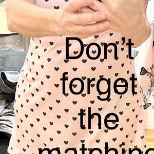 May include: A woman wearing a pink bucket hat with black hearts printed on it, a pink t-shirt with black hearts, and white sandals. The text "Don't forget the matching bucket hat" is superimposed on the image.