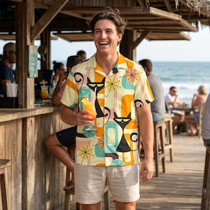 May include: A short-sleeved button-down shirt with a retro cat design in black, orange, and teal. The shirt is worn with beige shorts. The background shows a beach bar setting with people and the ocean.
