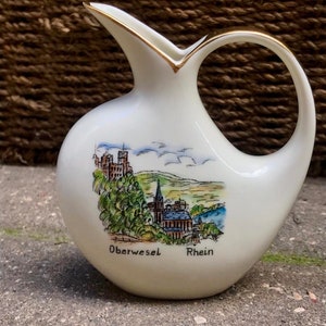 Op de afbeelding: Witte keramische kan met een gouden rand en een geschilderd tafereel van een stad met een kerk en de woorden "Oberwesel Rhein".