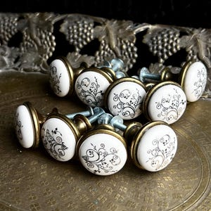 Könnte beinhalten: Ein Set runder Schrankknöpfe mit weißen Keramikoberflächen, schwarzen floralen Schnörkelmustern und antik messingfarbenen Metallsockeln. Jeder Knopf verfügt über eine Gewindeschraube, die zur Montage an der Rückseite herausragt.