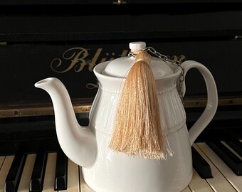 May include: A white ceramic teapot featuring a vertical ribbed pattern around the upper body and a matching lid. A light beige silk tassel is attached to the lid handle by a small silver-toned metal chain, draping over the front of the teapot.