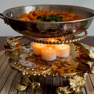 May include: A stainless steel food warmer pan containing cooked orange squash and herbs, resting on a gold-toned metal stand with ornate animal-shaped supports and a decorative base, illuminated by four small lit white tea light candles underneath.