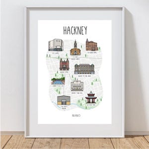 May include: A framed print featuring a map of Hackney, London, with illustrated landmarks like St. John's Hackney Church and Victoria Park. The word "HACKNEY" is at the top in black. The map is in a white frame.