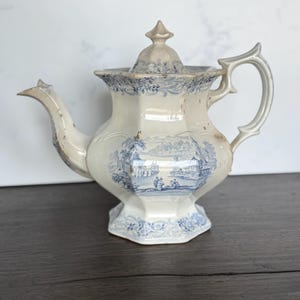 May include: Antique white and blue ceramic teapot with a landscape design. The teapot features a hexagonal base and body, a curved spout, and a decorative handle. The lid is topped with a pointed finial. The teapot is likely vintage.