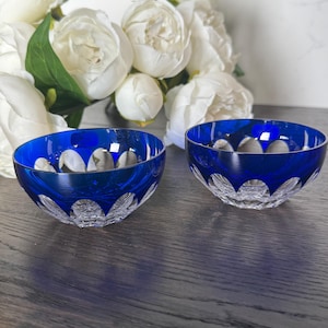 May include: Two small, round bowls crafted from deep blue glass with a clear, cut-glass base. Each bowl features a series of arched, scalloped cutouts around the exterior, revealing the transparent glass underneath the vibrant blue surface.