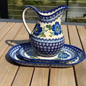 May include: A ceramic pitcher and matching oval tray featuring a cream-colored base with hand-painted blue flowers, green leaves, and intricate dark blue geometric patterns along the rims and base. The pitcher has a curved handle and spout.