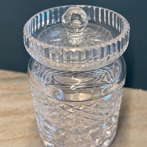 May include: A clear, cut-glass jar with a lid. The jar has a diamond pattern on the lower portion and vertical lines on the rim. The lid has a knob and a starburst design. The jar is sitting on a light-colored surface.