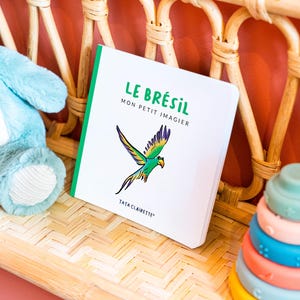 Peut inclure: Un livre pour enfants intitulé "LE BRÉSIL MON PETIT IMAGIER" avec une bordure verte et une illustration de perroquet coloré. Le livre est posé sur un plateau en bambou tressé, à côté d'un animal en peluche bleu et d'une pile d'anneaux colorés.