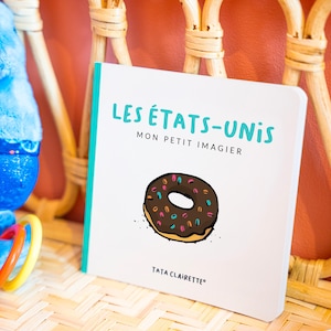 Peut inclure: Un livre cartonné carré à couverture blanche portant le texte "LES ÉTATS-UNIS" en bleu sarcelle, "MON PETIT IMAGIER" en noir et "TATA CLAIRETTE" en bas. Le centre de la couverture présente une illustration graphique d'un beignet au chocolat.