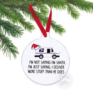 May include: A white, round Christmas ornament with a red ribbon, hanging from a green pine branch. The ornament features a postal truck wearing a Santa hat and the text: "I'M NOT SAYING I'M SANTA. I'M JUST SAYING, I DELIVER MORE STUFF THAN HE DOES."