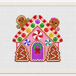 May include: A cross-stitch pattern on white Aida cloth featuring a pink gingerbread house decorated with colorful candy accents, a peppermint swirl, a green wreath on a purple door, and two smiling gingerbread figures peeking over the roof.