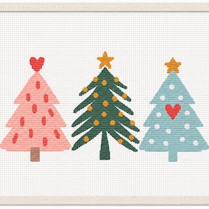 May include: A cross-stitch pattern on white Aida cloth featuring three stylized Christmas trees. The left tree is pink with red accents and a heart topper, the center is green with yellow ornaments and a star, and the right is light blue with white dots and a red heart.