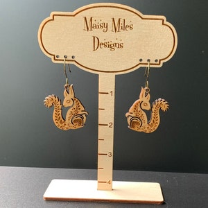 May include: A pair of wooden earrings featuring a squirrel design. The earrings are brown and have a detailed pattern. The earrings are hanging from a wooden display stand with the text "Maisy Miles Designs".
