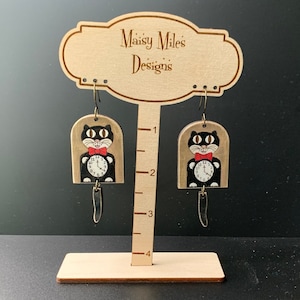 May include: A pair of dangle earrings featuring a black cat design with a red bow tie and a clock face. The earrings are displayed on a wooden stand with the text "Maisy Miles Designs".