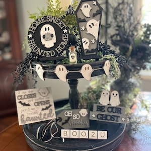 May include: A black tiered tray decorated with Halloween themed decorations. The tray features a sign that says "Home Sweet Haunted Home", a coffin with ghosts, a ghost garland, a sign that says "If one door closes and another one opens your house is probably haunted", a sign that says "Oh so spooky", a sign that says "Be our ghost", and a sign that says "Boo 2 U".
