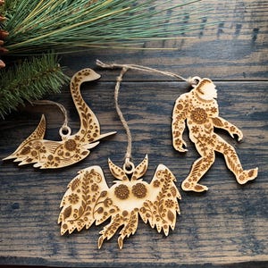 May include: Three wooden Christmas ornaments with intricate floral designs. The ornaments are shaped like a swan, a bird, and a Bigfoot.
