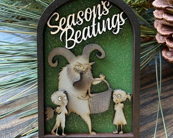 May include: A wooden Christmas ornament with a green background and the text "Seasons Beatings". The ornament features a cartoon monster with horns and a basket, and two children.