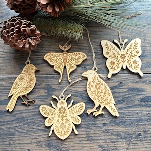 May include: Wooden ornaments in the shape of a bird, moth, butterfly, bee, and raven. Each ornament is intricately carved with floral designs and has a natural wood finish. They hang from twine strings, perfect for holiday decor.