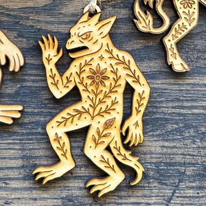 K&ouml;nnte beinhalten: Hellbraunes Holzornament mit einer stilisierten Kreatur mit floralem Design. Das Ornament zeigt eine detaillierte Schnitzerei einer monsterartigen Figur mit strengem Gesichtsausdruck und Krallenf&uuml;&szlig;en. Aufgeh&auml;ngt an nat&uuml;rlichem Bindfaden, erinnert es an Folklore.