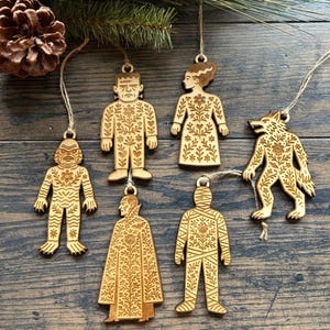 May include: Wooden Halloween ornaments featuring classic monster characters like Frankenstein, the Bride of Frankenstein, and a werewolf. Each ornament is intricately carved with floral patterns and includes a string for hanging. The ornaments are made of light-colored wood.