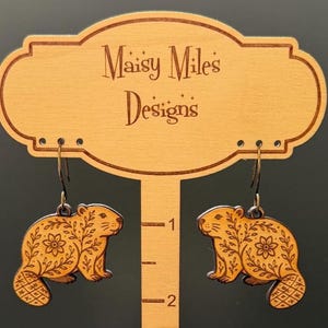May include: A pair of wooden earrings featuring a beaver design with floral and geometric patterns. The earrings are displayed on a wooden stand with the text "Maisy Miles Designs". The earrings are approximately 1 inch in length.