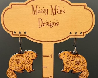 Könnte beinhalten: Holzohrringe mit einem Biber-Design und floralen und geometrischen Mustern. Die Ohrringe werden auf einem Holzständer mit dem Text "Maisy Miles Designs" präsentiert. Die Ohrringe sind etwa 2,5 cm lang.
