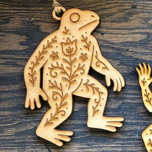 K&ouml;nnte beinhalten: Holzornament in Froschform mit aufwendigen Blumen- und Blattschnitzereien. Der Frosch ist hellbraun mit nat&uuml;rlicher Holzmaserung. Zum Aufh&auml;ngen ist eine Juteschnur befestigt. Das Design zeigt einen stilisierten Frosch mit detaillierten botanischen Mustern.