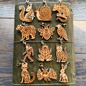 May include: A collection of wooden animal charms, including a squirrel, snail, bear, bee, bird, bigfoot, frog, cat, fox, bat, and rabbit. Each charm is intricately carved with floral patterns and has a metal clasp for attachment.