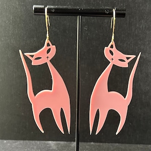 May include: A pair of pink cat-shaped earrings with gold hooks. The cats are stylized and have a simple design.