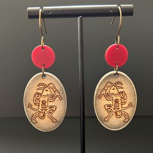 May include: A pair of wooden earrings with a red circle and a detailed wood carving of a goat-like figure. The earrings are hanging from a black stand.