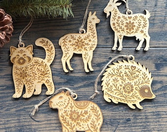 Puede incluir: Conjunto de cinco adornos de animales de madera, incluyendo un panda rojo, una llama, una cabra, un erizo y un carpincho. Cada adorno está intrincadamente tallado con patrones florales y de hojas y tiene una cuerda de yute para colgar. Se exhiben sobre madera oscura.