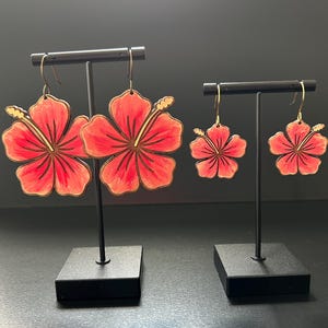 May include: Two pairs of hibiscus flower earrings, one pair is larger than the other. The earrings are made of wood and painted in shades of pink and yellow.