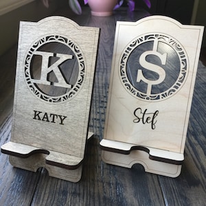 May include: Two wooden phone stands with a decorative design. The stand on the left has a K in a circle and the name Katy. The stand on the right has a dollar sign in a circle and the name Stef.