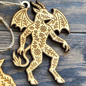 K&ouml;nnte beinhalten: Holzornament mit einer geh&ouml;rnten, gefl&uuml;gelten Figur mit floralen Details. Das Ornament aus hellem Holz hat eine Aufh&auml;ngeschlaufe. Das Design ist aufwendig und detailliert, mit einer rustikalen &Auml;sthetik. Es ist etwa 10 cm hoch.