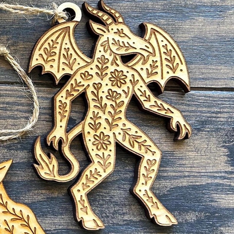 K&ouml;nnte beinhalten: Holzornament mit einer geh&ouml;rnten, gefl&uuml;gelten Figur mit floralen Details. Das Ornament aus hellem Holz hat eine Aufh&auml;ngeschlaufe. Das Design ist aufwendig und detailliert, mit einer rustikalen &Auml;sthetik. Es ist etwa 10 cm hoch.