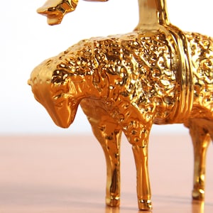 May include: A gold-colored metal figurine of a lamb with a textured surface. The lamb is standing on a wooden surface.