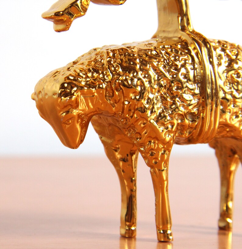 May include: A gold-colored metal figurine of a lamb with a textured surface. The lamb is standing on a wooden surface.